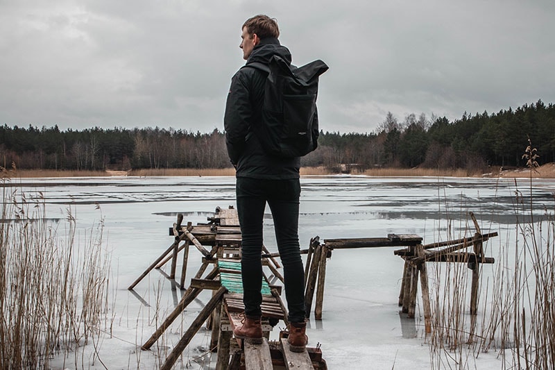 A man with black jeans and a backpack standing on a wooden pier near a frozen lake